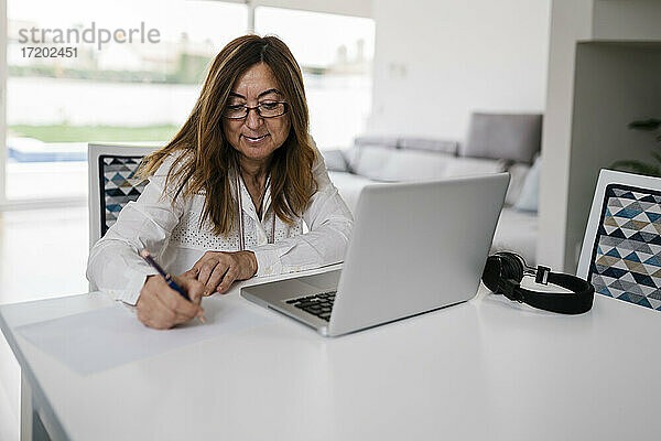 Berufstätige Frau  die mit Laptop und Kopfhörern zu Hause sitzt und auf Papier schreibt