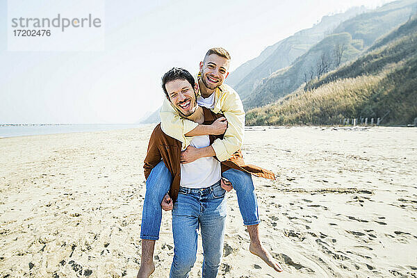 Fröhlicher schwuler Mann  der seinen Freund im Urlaub am Strand huckepack nimmt