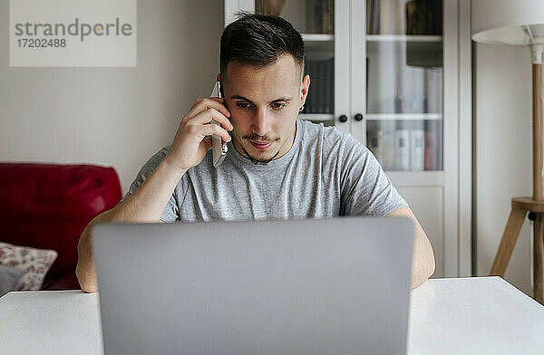 Junger männlicher Unternehmer  der mit seinem Smartphone spricht  während er im Büro zu Hause einen Laptop benutzt