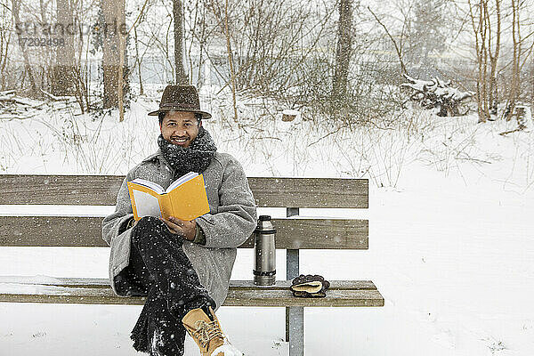 Lächelnder Mann liest ein Buch  während er im Winter auf einer Bank sitzt