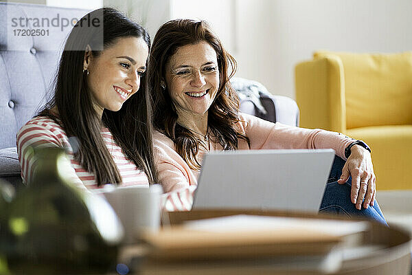 Lächelnde Mutter und Tochter schauen gemeinsam auf den Laptop  während sie zu Hause im Wohnzimmer sitzen