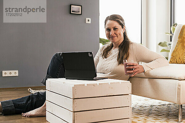 Lächelnde Frau mit einem Glas Saft und einem digitalen Tablet im Wohnzimmer sitzend