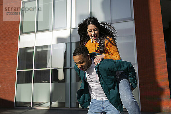Mann nimmt seine fröhliche Freundin vor einem modernen Gebäude huckepack