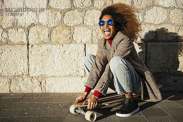 Fröhliche Frau mit Sonnenbrille  die auf einem Skateboard an der Wand sitzt  an einem sonnigen Tag