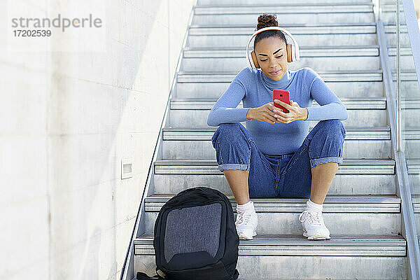 Junge Frau mit Rucksack und Kopfhörern  die auf einer Treppe sitzend ein Mobiltelefon benutzt