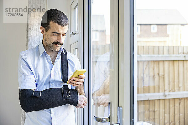 Mann mit Armschlinge  der sein Smartphone benutzt  während er zu Hause am Fenster steht