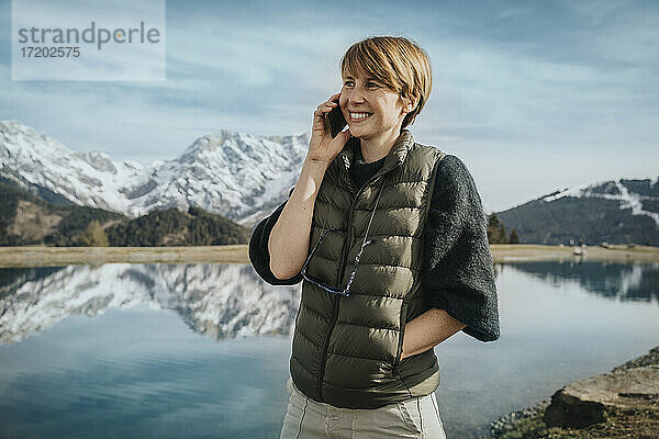 Lächelnde Frau mit Hand in der Tasche  die mit ihrem Handy telefoniert und dabei gegen den Himmel am Prinzensee  Hochkönigmassiv  Salzburger Land  Österreich  blickt