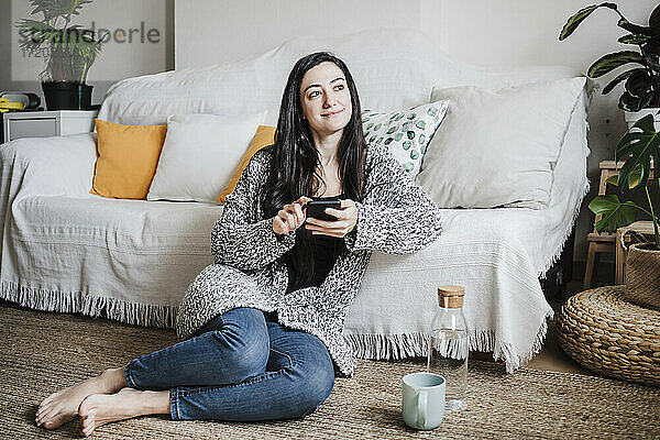Lächelnde Frau schaut weg  während sie ihr Smartphone auf dem Sofa im Wohnzimmer benutzt