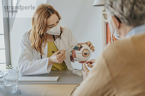 Augenärztin mit Gesichtsmaske erklärt dem Patienten in der Klinik sitzend ein Augenmodell