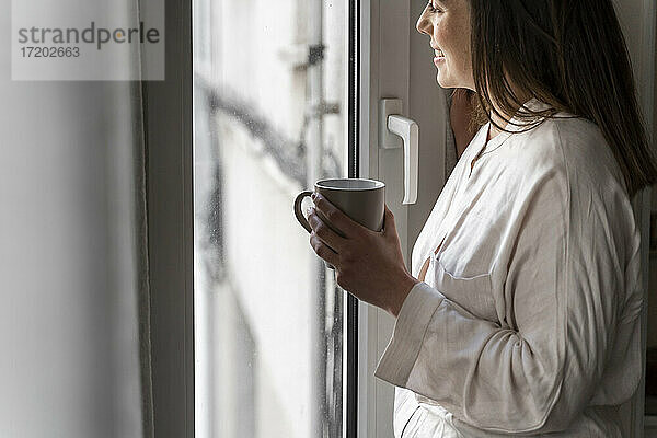 Glückliche junge Frau mit Kaffeetasse  die durch das Fenster ihres Hauses schaut