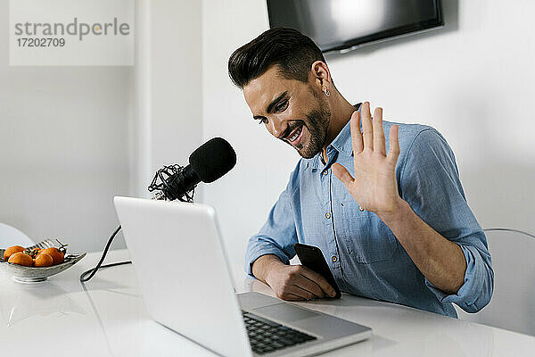 Lächelnder Geschäftsmann mit winkender Hand am Laptop  während er zu Hause sitzt