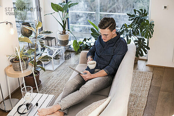Junger Mann mit Kaffeetasse auf dem Sofa sitzend und mit digitalem Tablet im Wohnzimmer