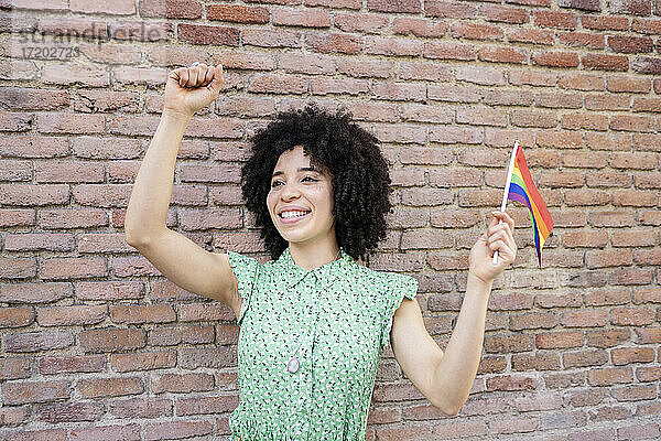 Fröhliche junge Frau ballt die Faust  während sie die LGBTQI-Regenbogenfahne gegen eine Backsteinmauer hält