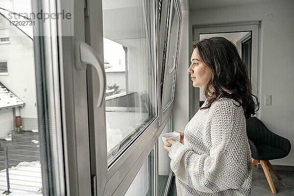 Frau steht mit Kaffeetasse und schaut durch das Fenster zu Hause