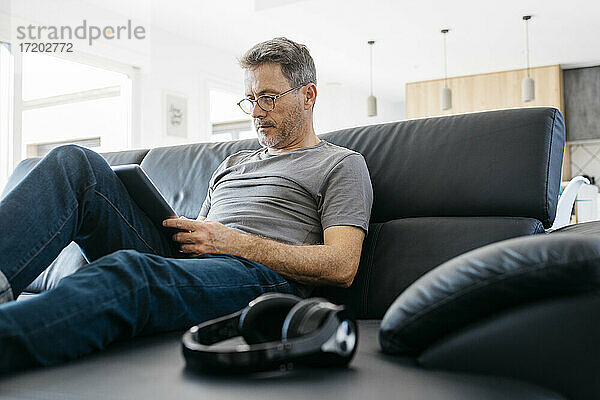 Älterer Mann  der ein digitales Tablet benutzt  während er auf dem Sofa im Wohnzimmer sitzt