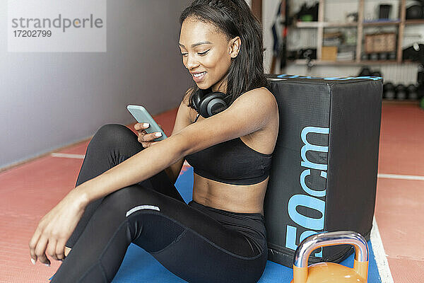 Lächelnde junge Frau  die sich an eine Box lehnt und ein Mobiltelefon im Fitnessstudio benutzt