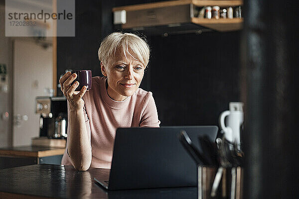 Lächelnde Geschäftsfrau  die auf einen Laptop schaut  während sie eine Kaffeetasse am Küchentisch im Heimbüro hält