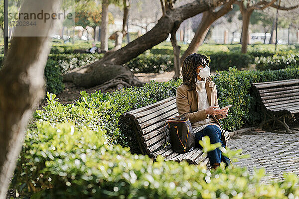 Ältere Frau mit Schutzmaske  die ein Handy hält  während sie im Park sitzt