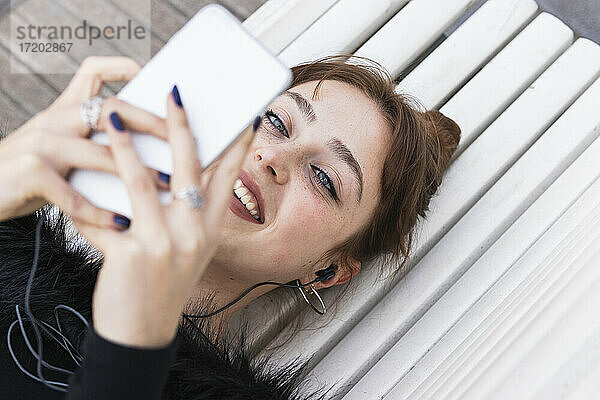 Lächelnde junge Frau  die auf einer Bank liegend im Internet surft
