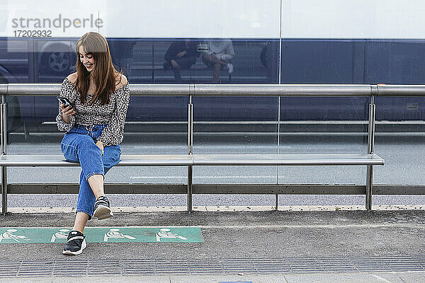 Lächelnde junge Frau  die ein Smartphone benutzt  während sie auf einer Bank an einer Straßenbahnhaltestelle sitzt