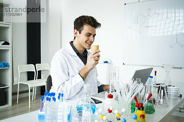 Lächelnder männlicher Wissenschaftler  der einen Kaffee trinkt  während er in einem Labor am Laptop arbeitet