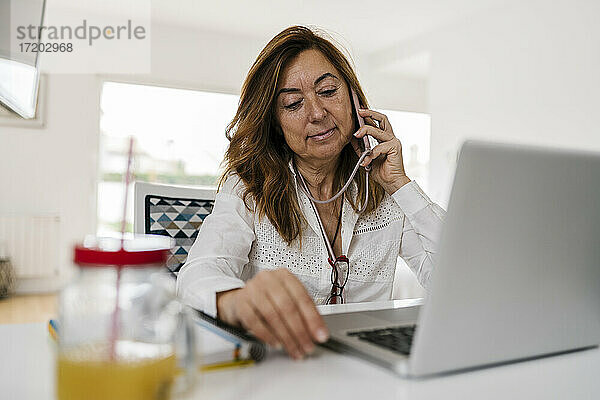 Unternehmerin  die mit ihrem Smartphone telefoniert  während sie zu Hause mit ihrem Laptop auf dem Schreibtisch sitzt
