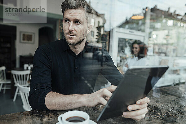 Geschäftsmann  der ein digitales Tablet benutzt und wegschaut  während er in einem Café sitzt