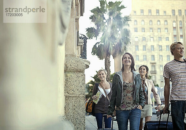 Lächelnde männliche und weibliche Freunde mit Koffern  die auf dem Gehweg einer Straße in der Stadt spazieren gehen