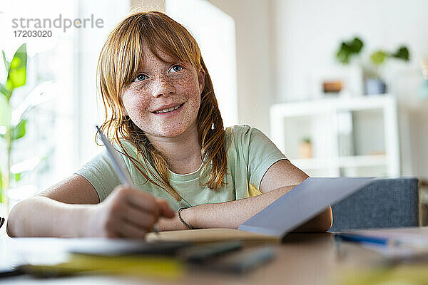 Nettes lächelndes rothaariges Mädchen  das wegschaut  während es zu Hause Hausaufgaben macht