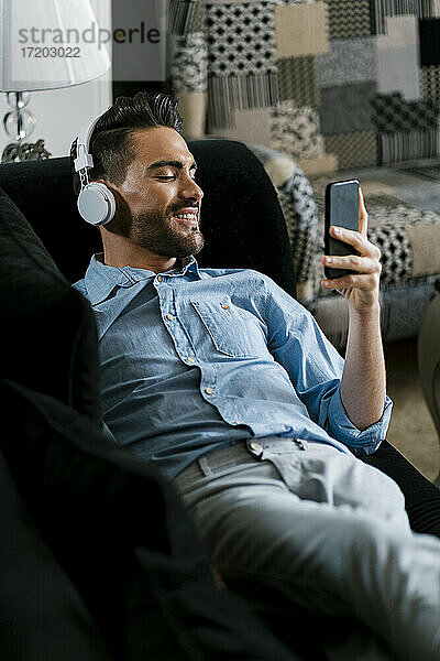 Lächelnder Mann mit Kopfhörern  der ein Mobiltelefon benutzt  während er zu Hause auf dem Sofa liegt