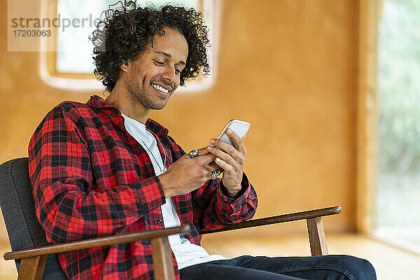 Junger Mann  der ein Smartphone benutzt  während er in einem geräumigen Zimmer auf einem Sessel sitzt