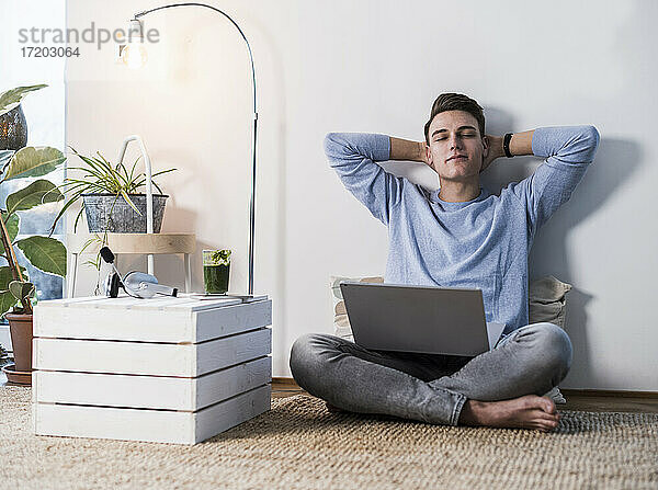 Junger Mann mit Laptop sitzt entspannt an der Wand im Wohnzimmer