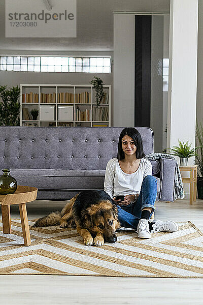 Lächelnde Frau sitzt mit Hund auf Teppich vor Sofa im Wohnzimmer