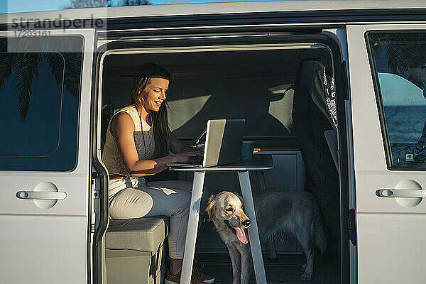 Lächelnde Frau arbeitet am Laptop im Wohnmobil an einem sonnigen Tag am Wochenende