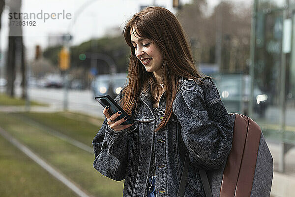 Lächelnde Frau  die ein Smartphone benutzt  während sie an einer Straßenbahnhaltestelle steht