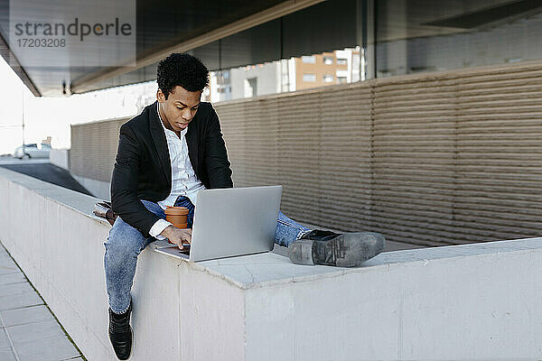 Mittlerer erwachsener Geschäftsmann  der einen Laptop benutzt  während er auf einer Stützmauer sitzt