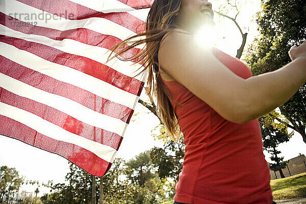 Junge Frau läuft im Park  während sie die amerikanische Flagge an einem sonnigen Tag hält