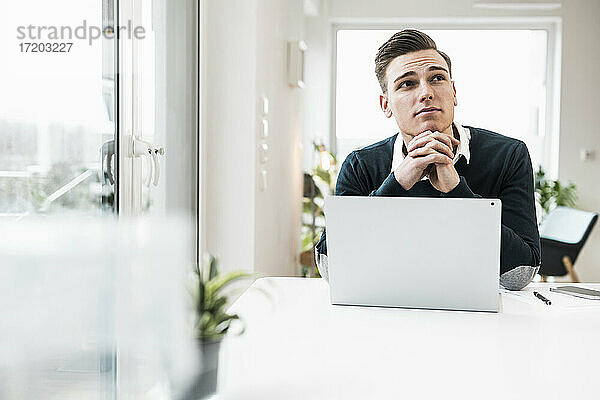 Nachdenklicher männlicher Unternehmer mit Laptop  der am Schreibtisch sitzt und wegschaut