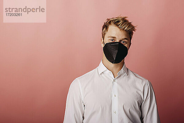 Junger Geschäftsmann mit Gesichtsschutzmaske  der vor einem farbigen Hintergrund steht und starrt