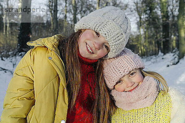 Lächelnde Schwestern in warmer Kleidung  die zusammen gegen Bäume im Schnee stehen