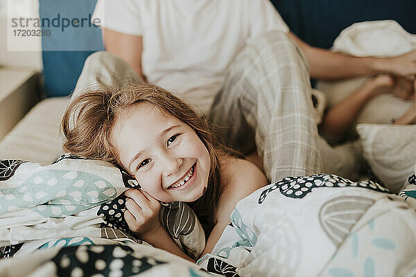 Fröhliches Mädchen auf dem Bett liegend mit Vater im Hintergrund sitzend