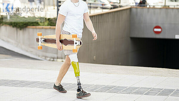Junger behinderter Mann hält Skateboard  während er auf dem Fußweg geht