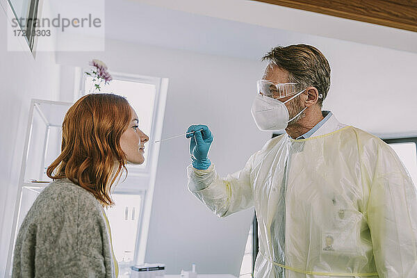 Arzt in Schutzanzug und Gesichtsmaske bei der Untersuchung eines Patienten im Untersuchungsraum