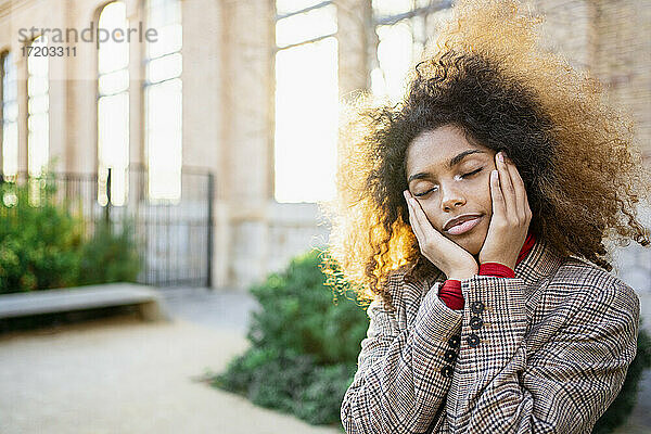 Afro-Frau mit braunem Haar steht mit geschlossenen Augen vor einem Gebäude