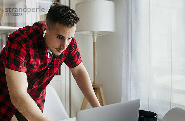 Männlicher Unternehmer mit In-Ear-Kopfhörern  der sich auf einen Tisch stützt  während er im Büro zu Hause einen Laptop benutzt