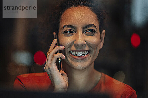 Junge Frau lächelt  während sie in einem Café sitzt und mit ihrem Handy spricht