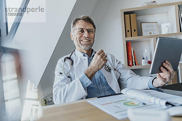 Lächelnder männlicher Arzt mit digitalem Tablet  der am Schreibtisch in einer Arztpraxis sitzt
