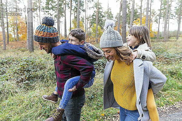 Lächelnde Eltern  die ihre Kinder beim Spaziergang im Wald huckepack nehmen