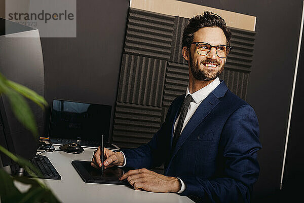 Lächelnder Geschäftsmann schaut weg  während er ein Grafiktablett am Schreibtisch im Büro benutzt
