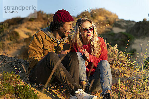 Freundin lächelt  während sie neben ihrem fröhlichen Freund auf einer Sanddüne bei Sonnenuntergang sitzt